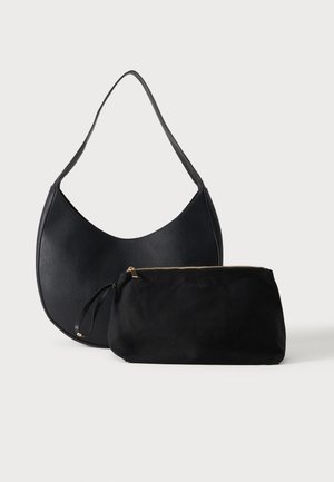 Bolso de hombro de cuero negro con correa ancha y bolsa de tela negra con cremallera colocada al frente sobre un fondo claro y liso.