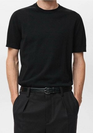 Homme portant une chemise noire unie à manches courtes rentrée dans un pantalon noir plissé avec une ceinture noire, debout, les mains dans les poches.
