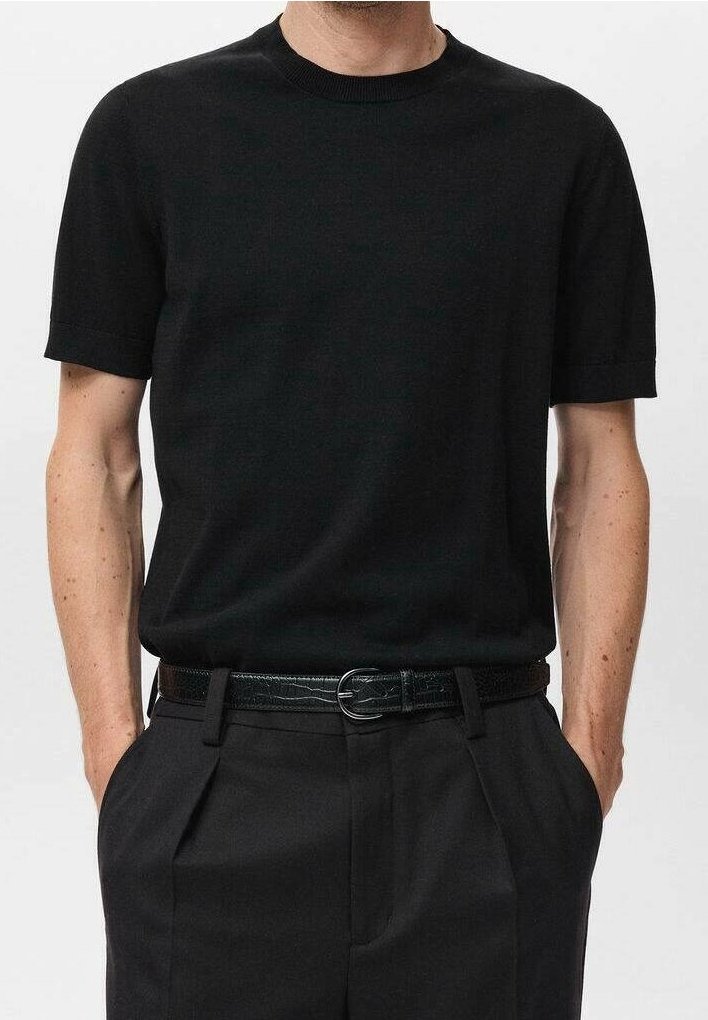 Homme portant une chemise noire unie à manches courtes rentrée dans un pantalon noir plissé avec une ceinture noire, debout, les mains dans les poches.