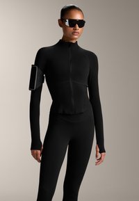 Schwarze sportliche Zippjacke mit langen Ärmeln, glatter Textur und geschnittenem Design, kombiniert mit Leggings und dunklen Sonnenbrillen. Armband für das Gerät inklusive.