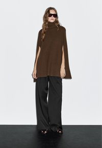 Bruine geribbelde turtleneck poncho met zijsplitten, gecombineerd met zwarte wijde broek en zwarte open sandalen. Model draagt een zonnebril.