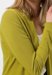 Hellgrüner, strukturierter Cardigan mit Wabenmuster, offenem Vorderdesign und langen Ärmeln, kombiniert mit einem weißen Top darunter.