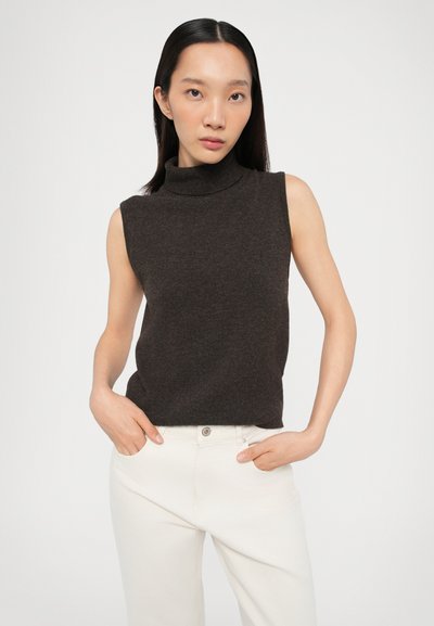 Smeđa bez rukava turtleneck pulover u mekom, teksturiranom pletivu. Uparen s bijelim hlačama visokog struka. Model stoji s rukama na bokovima.