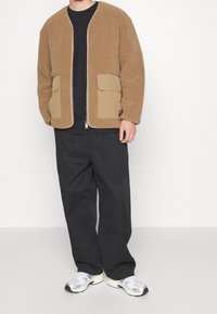 Brązowa, puszysta kurtka polarowa z zamkiem i dwoma przednimi kieszeniami, zestawiona z czarnymi spodniami i szarymi sneakersami. Prosty design z luźnym krojem.