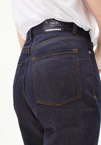 Jean en denim bleu foncé avec des surpiqûres oranges, comportant une poche arrière et une étiquette au niveau de la taille. Texture lisse et design ajusté.