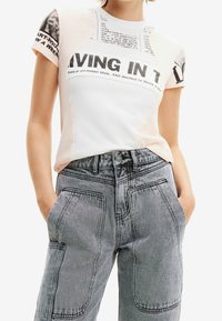 Camiseta blanca con estampado de periódico, acentos rosas, cuello redondo, combinada con unos jeans grises de tiro alto que presentan grandes bolsillos de parche y detalles de costura.