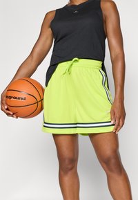  atletisk sort tanktop, neongrøn shorts med sort og hvid striber, elastisk talje, parret med en orange basketball.