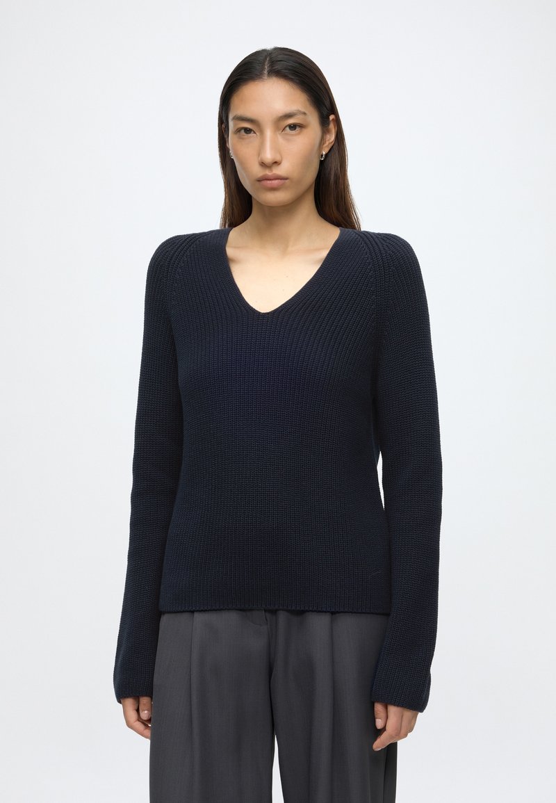 Donna con i capelli lunghi e scuri che indossa un maglione a maglia blu navy scuro con scollo a V e pantaloni a gamba larga grigio scuro su uno sfondo bianco semplice.