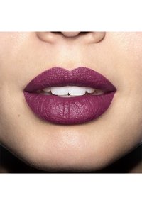 Matte pruimenlipstick aangebracht op vollere lippen, met zichtbare textuur, en een contrasterende witte glimlach eronder. De huidskleur is licht.