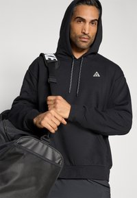 Μαύρο φούτερ με κουκούλα και τρίγωνο λογότυπο, κορδόνια και ριμπ μανσέτες, φορεμένο με μαύρη τσάντα που διαθέτει διχτυωτή επιφάνεια.