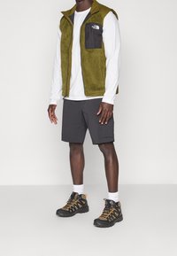 Grön fleeceväst med en svart ficka, vit långärmad skjorta, grå shorts och svarta vandringsskor med orange detaljer.