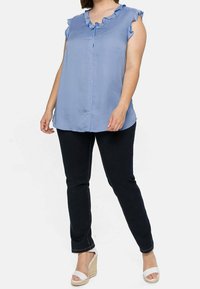 Blusa sin mangas de color azul claro con cuello con volantes, frontal abotonado, combinada con pantalones de mezclilla oscuros y cuñas blancas de plataforma.