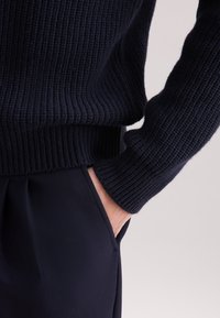 Granatowy sweter z prążkowanym splotem z dopasowanym pasem i mankietami, noszony na ciemnych spodniach, podkreślający gładką fakturę tkaniny.