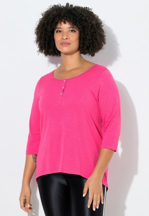 Frau mit lockigem Haar, die ein knallpinkes, geknöpftes Dreiviertelarm-Oberteil und schwarze Leggings trägt, steht vor einem hellgrauen Hintergrund.