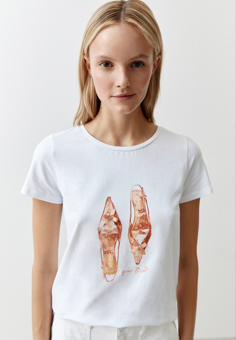 White cotton t-shirt featuring an illustration of two high-heeled shoes, detailed with bows and accents in shades of orange and pink.