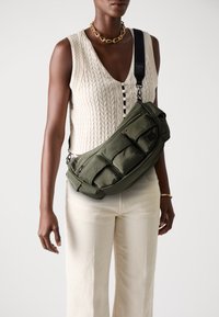 Sac en nylon vert avec plusieurs poches zippées, texture lisse et bandoulière réglable, porté sur un haut en tricot blanc et un pantalon beige.