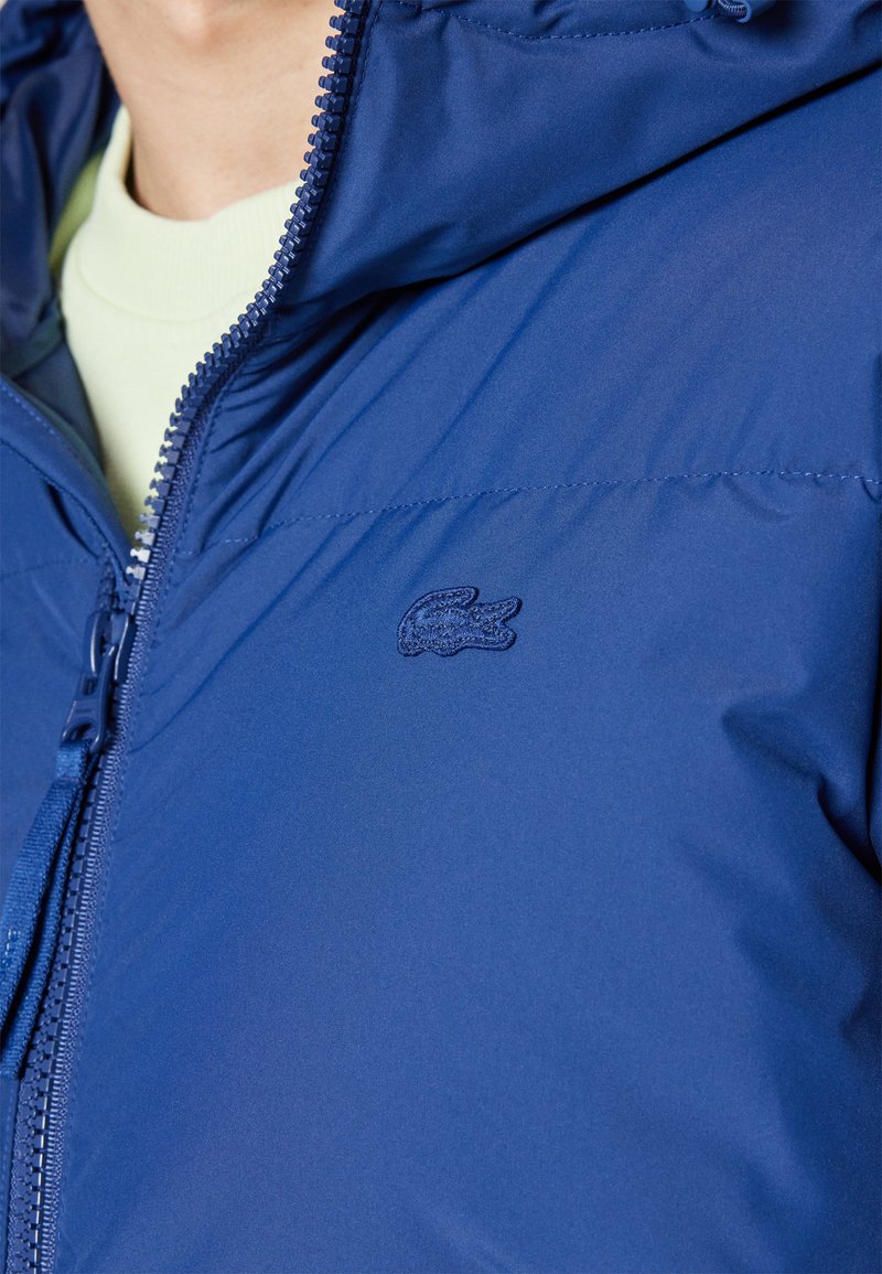 Chaqueta de plumas azul con un material liso y resistente al agua, cierre con cremallera y un logo bordado de Lacoste en el pecho. Cuenta con capucha.