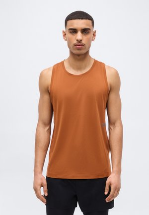 Νέος άνδρας με κοντά μαλλιά που φοράει αμάνικο τοπ χρώματος σκουριάς και μαύρο σορτς, στέκεται μπροστά σε απλό ανοιχτό φόντο.