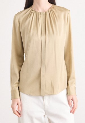 Kvinde iført en beige bluse med lange ærmer med sammenrullet halsudskæring og hvide bukser, stående mod en ensartet lys baggrund.