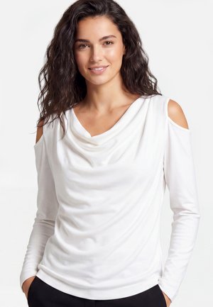 Donna con capelli lunghi e scuri che indossa una maglia bianca a maniche lunghe con spalle scoperte e pantaloni neri, sorridente con le mani nelle tasche.