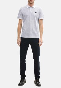 Polo blanc avec un motif bleu subtil, présentant un col classique et un petit logo sur la poitrine. Associé à un pantalon sombre.