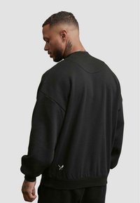 Černá mikina s kulatým výstřihem, volným střihem a texturovanou tkaninou. Obsahuje spadlé ramena a malou logo aplikaci blízko spodního lemu.