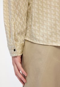 Langarmshirt van beige stof met een geometrisch patroon, met een knoopsluiting bij de manchetten en een gladde textuur. Gepaard met beige broeken.