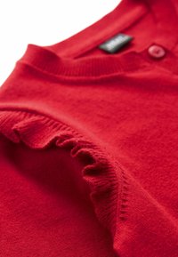 Gros plan d'un vêtement en maille rouge avec bord de manche à volants et bouton près du col, montrant la texture douce du tissu et les coutures fines.