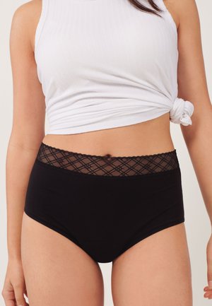 Žena nosiace čierne nohavičky s vysokým pásom s čipkovaným detailom a biele tielko bez rukávov so zaväzovaním, stojaca pred jednoduchým pozadím.