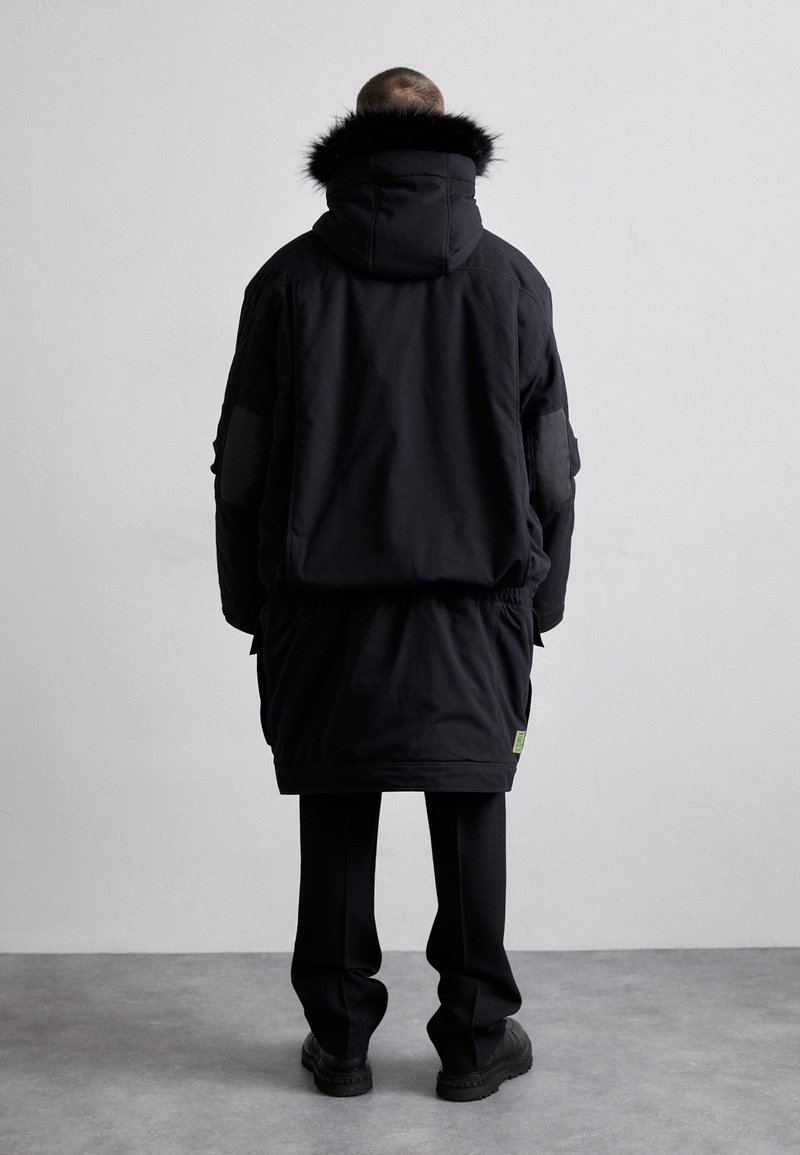 Schwarze Parka mit einem gefüttert mit Fell, langem Design, elastischem Bund und seitlichen Taschen. Kombiniert mit schwarzen Hosen und klobigen schwarzen Stiefeln.