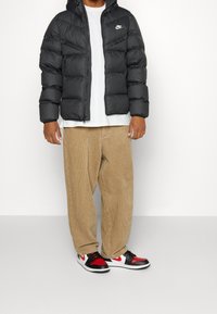 Black puffer jacket with a hood and white logo, worn over a white t-shirt, paired with beige corduroy pants and red, black, and white sneakers.