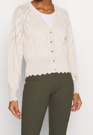 Žena oblečená v béžovém pleteném svetru na knoflíky se vzorem cikcak a zakončeném vroubkováním, doplněném olivově zelenými žebrovanými kalhotami.