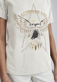 Beige T-Shirt mit kurzen Ärmeln, das ein Grafikdruck eines Adlers mit goldenen Akzenten und einem Sternenhintergrund zeigt, aus weichem Baumwollmaterial gefertigt.