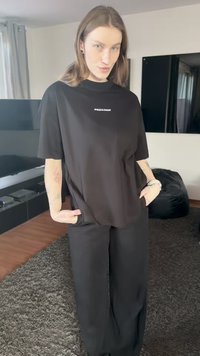 Black oversized t-shirt with short sleeves and a round neckline features a white logo in the center. Paired with loose black trousers.