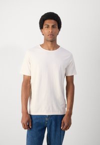 Cămașă din bumbac bej deschis, cu mâneci scurte și guler rotund. Croială lejeră cu o textură subtilă și detalii de cusătură. Asortată cu blugi din denim albastru.