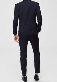 Mies, joka on pukeutunut istuvaan mustaan pukuun ja mustiin pukukengiin, seisoo yksi käsi taskussa, kuvattuna selkäpuolelta yksinkertaista taustaa vasten.