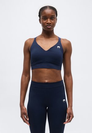 Γυναίκα που φοράει ασορτί αθλητικό σουτιέν και κολάν σε σκούρο μπλε χρώμα με λευκό λογότυπο στο στήθος και στο μηρό, στέκεται μπροστά σε απλό φόντο.