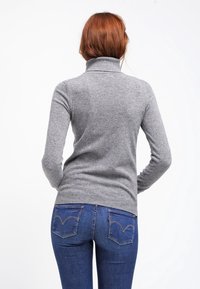 Kvinna med röd hår som står vänd bort, iklädd en grå höghalset tröja och blå jeans med bakfickor synliga.