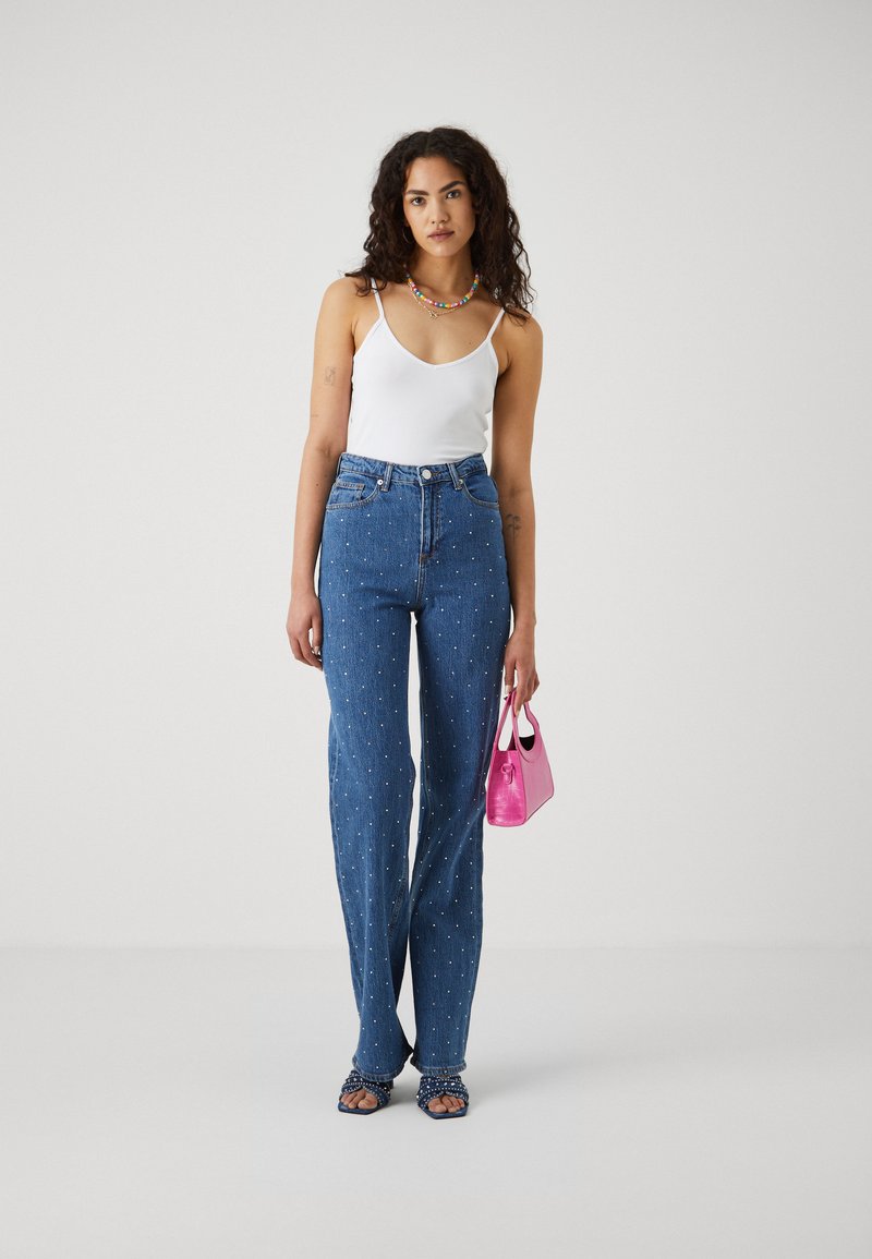 Blå denim jeans med perlebroderinger, kombinert med en hvit tanktop, en rosa håndveske og sandaler med åpen tå og rynkede stropper.