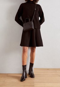 Pull noir oversize avec col haut, jupe noire évasée, bottines noires en cuir avec talons carrés, et un petit sac bandoulière noir.