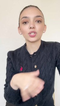 Cardigan preto em ponto de malha com botões de latão e um logótipo vermelho. Gestos individuais com as mãos, tom de pele claro, fundo com iluminação suave.