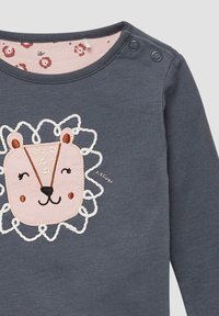 Graues Langarmshirt mit einem Löwenkopf-Design in Pink, verziert mit weißen Ziernähten, silbernen Druckknöpfen an der Schulter und einem rosa gemusterten Kragen.