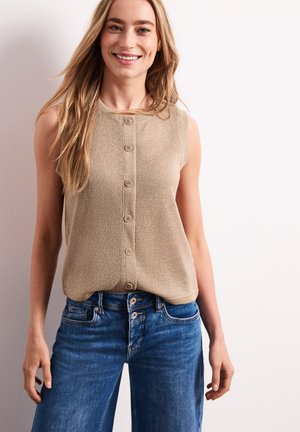 Femme souriante aux longs cheveux blonds portant un haut beige sans manches à boutons et un jean bleu, sur un fond clair uni.