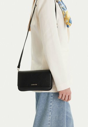 Osoba ubrana w beżową marynarkę i niebieskie dżinsy niesie czarną torebkę na ramię marki Lasocki z długim paskiem.