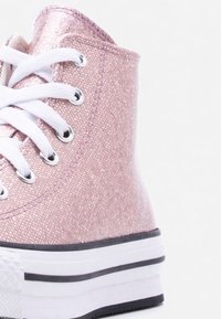 Αθλητικό παπούτσι τύπου high-top με ροζ υφασμάτινο ύφασμα με γκλίτερ, λευκά κορδόνια, ασημί μεταλλικά τρύπες και λευκή σόλα με μαύρες ρίγες, εμφανιζόμενο από την πλαϊνή και πίσω γωνία.