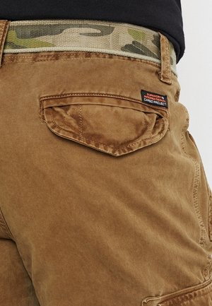 Brun cargobukselomme med klap og Superdry International Cargo Project-label, båret med et camouflagemønster bælte og sort top.