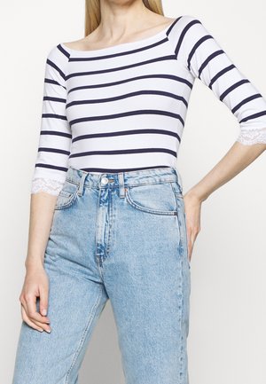 Vrouw die een wit en marineblauw gestreepte boothals top draagt met mouwen afgezet met kant en een high-waisted lichtblauwe spijkerbroek.