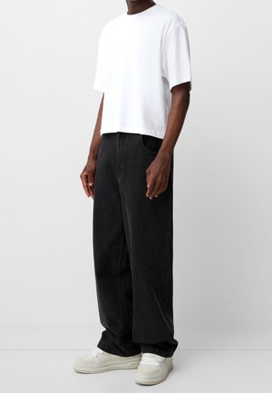 Man draagt losse zwarte jeans, een wit cropped t-shirt en witte sneakers, staand tegen een effen witte achtergrond.