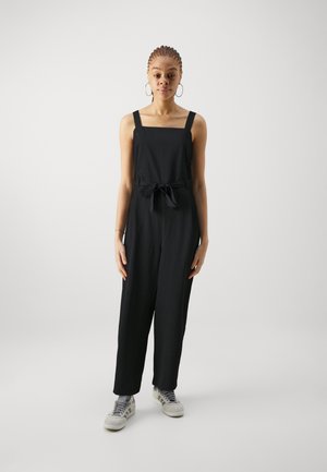 Mono negro con escote cuadrado, tirantes ajustables y cintura anudada. La modelo lleva zapatillas grises y está de pie sobre un fondo liso.