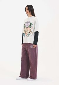 Witte oversized graphic t-shirt met een bloemenontwerp en tekst, gecombineerd met losse bordeauxrode broeken en witte sneakers.